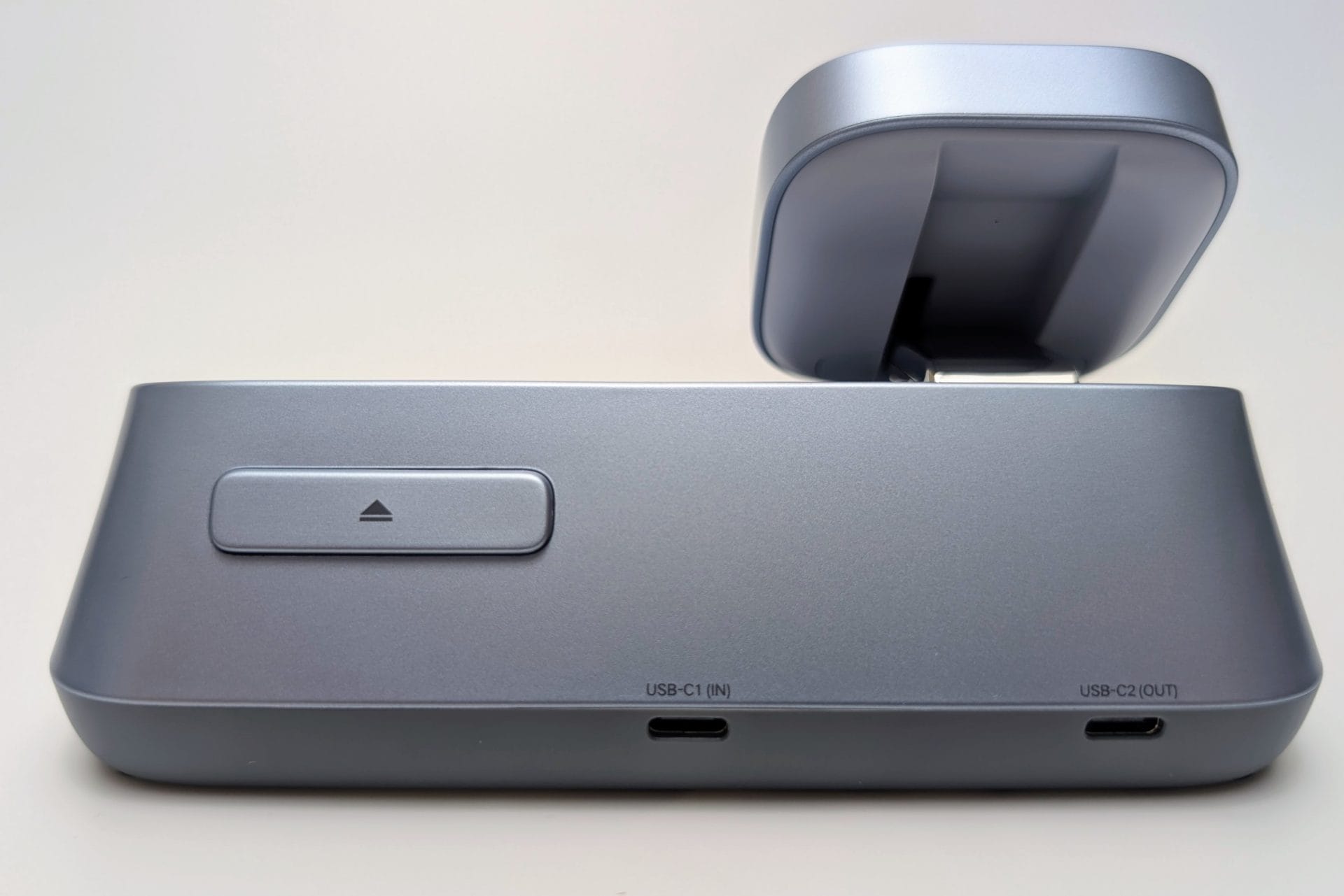 UGREEN MagFlow 3-in-1 Magnetic Wireless Desktop Charger A side view of the UGREEN MagFlow 3-in-1 Magnetic Wireless Charger, focusing on the rear ports and the button for deploying the Apple Watch charger. The main unit is a smooth, light grey plastic. On the back, there is a horizontal button with a small arrow pointing up, which is used to release the Apple Watch charging arm. Below, there are two USB-C ports: one labeled "USB-C1 (IN)" for power input, and one labeled "USB-C2 (OUT)" for charging an additional device with a cable. The main MagSafe-compatible phone charger is visible in the raised position at the top.