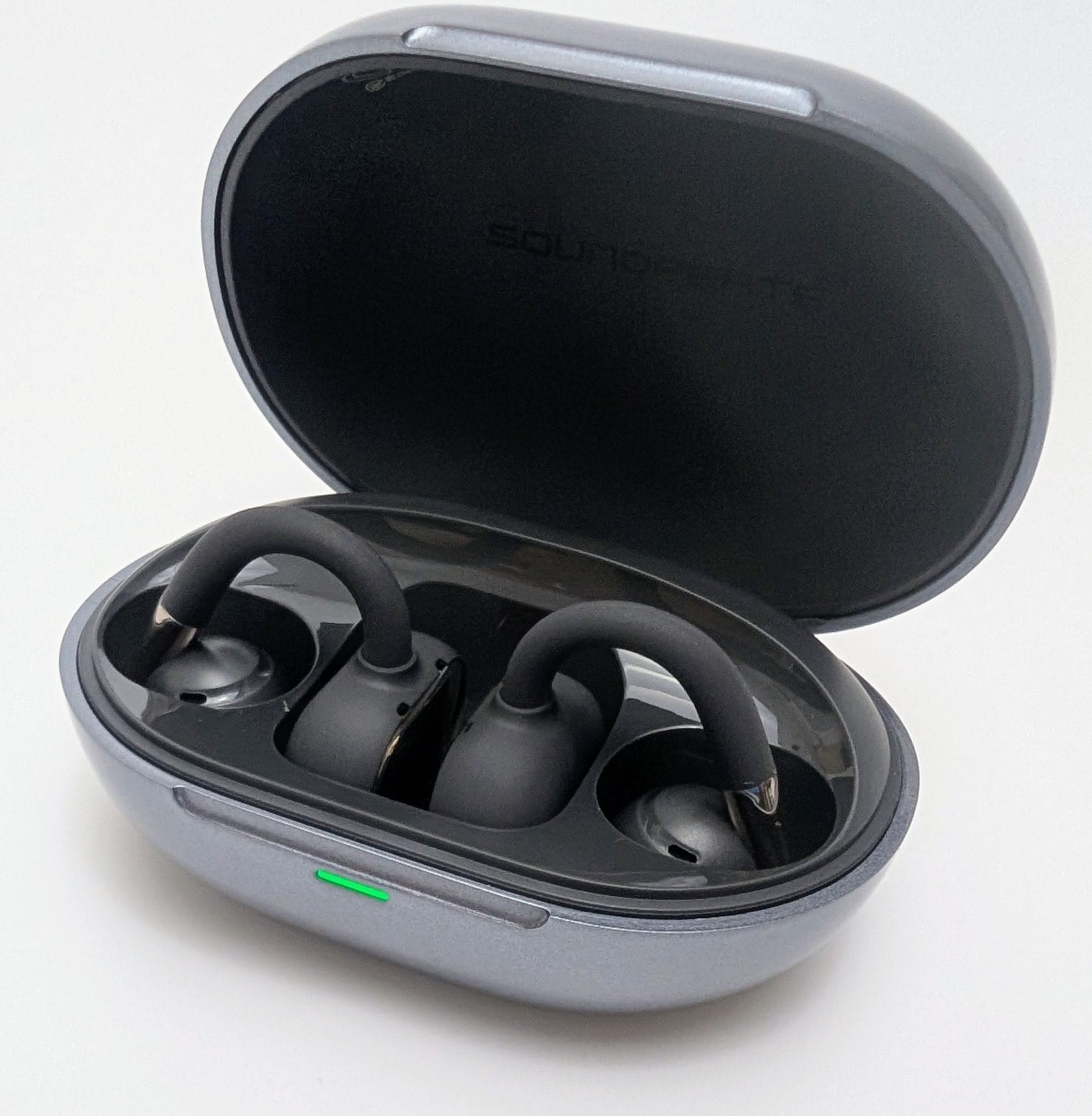 Open charging case of the SoundPEATS Clip1 wireless earbuds in metallic gunmetal gray. The lid is flipped back, revealing the two matte black open-ear clip-style earbuds securely seated in their molded slots with charging contacts aligned. A small green LED indicator glows on the front edge of the case, showing it is powered or charging. The interior lid has the faint embossed text "SOUNDPEATS" near the hinge. Photographed against a plain white background.